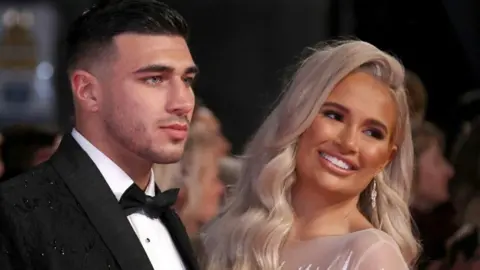 Getty Images Tommy Fury, wearing a black tuxedo with white shirt and black bow-tie, and Molly-Mae Hague, wearing a sheer dress and with long blonde hair, looking off c amera - Molly-Mae is smiling