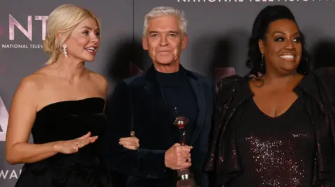 Getty Holly Willoughby, wearing a black strapless evening dress, stands with Philip Schofield, centre and holding an award, and Alison Hammond, right, wearing a maroon glittery dress and jacket