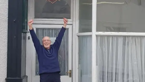 Dorothy Lambert Charlie Lambert celebrating outside Norman Nicholson's house after the property was bought in February. Mr Lambert is smiling and his hands are up in the air. He is wearing a navy blue sweatshirt and has glasses. The house has large front windows  with white panes. 