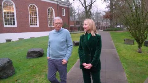 Maxine Collins/BBC Bill Gates in pale blue jumper and grey trousers and Katie Razzall in velvet green trouser suit walking outside Lakeside School in Seattle