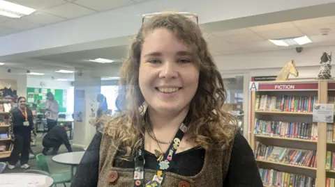 BBC/Kit Taylor A white young woman with curly brown hair. She is wearing a black top and brown dress and smiling.
