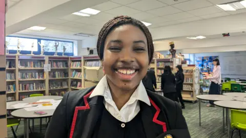 BBC/Kit Taylor A Nigerian teenage girl with brown braided hair and school uniform. She is smiling.
