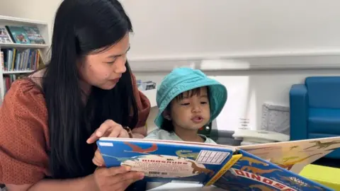 Andrew Turner/BBC A mother and her daughter read a book. The mother has long black hair, and is wearing a brown/rust coloured top. She is holding a book  that her young daughter appears to be reading. The child has shoulder length dark hair, and is wearing a white top and a turquoise sun hat. There is a book in the foreground, with the word "dinosaur" visible. A blue sofa is to the centre right of the image, and book shelves to the left.