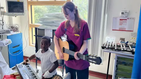 Ollie Conopo/BBC Woman with long brown hair in purple top playing a classical guitar with a young boy in a white T-shirt playing a keyboard. They are in a room with white walls and a large window,. There is medical equipment to the left. There is a pipe gamelan on a stand to the right.