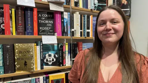 Jo Burn/BBC A woman with long, brown hair looking at the camera. She is wearing an orange/brown dress and is standing in front of a full bookshelf.