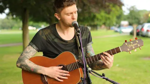 BBC A young man with short brown hair and tattooed arms is stood in a green area holding a brown acoustic guitar and singing into a microphone.