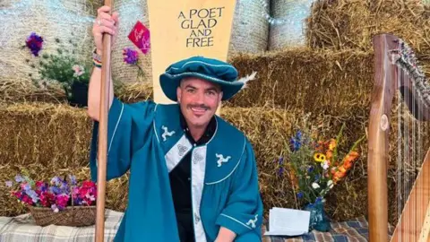 Team Bard Bradley Chambers wears a blue gown with a flamboyant traditional hat of a bard and holds a staff, in front of a sign that reads a poet glad and free. A harp and hay bales can be seen in the background. 