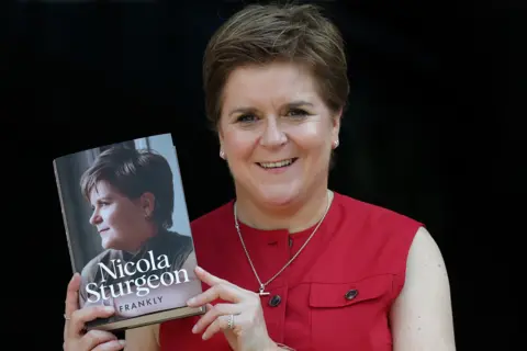 Getty Images Nicola Sturgeon, who has short brown hair combed in a side shed and is wearing a sleeveless red blouse, holding a copy of her memoir, Frankly.