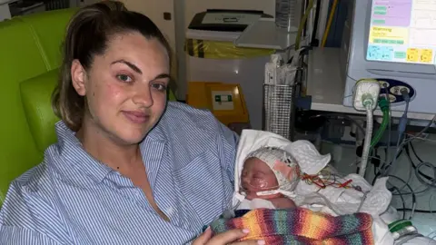 Olivia Dewar A woman with blonde hair, wearing a blue and white striped shirt, holds a newborn baby wrapped in a multicoloured blanket. There are tubes connecting the baby girl to a machine.