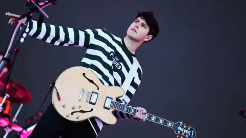 Vampire Weekend's guitarist on stage. He is wearing a black and white striped top and is holding a pale wood guitar.