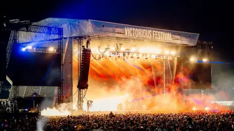 Victorious Festival Victorious Festival's main stage fully lit up with smoke and strobe lights. A sea of people are standing in front of the stage.
