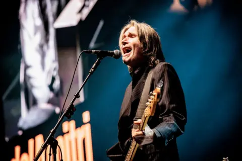 Getty Images Justin Currie is holding a guitar and singing into a microphone.