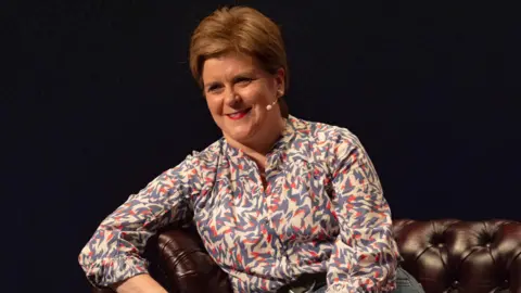 PA Media Nicola Sturgeon sits on a brown leather Chesterfield sofa. She is wearing a patterned blouse and jeans. She has a small microphone attached to her head.