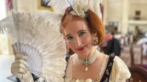 BBC Jane Austen tour guide Theresa Roche is dressed in cream and purple Georgian clothing with g loves, a pearl necklace and a feathery head piece. She is holding an open fan in her right hand and she is smiling.