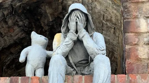 BBC A close up view of a grey sculpture depicting a seated, hooded male figure with hands covering its face, accompanied by a bear-like companion offering comfort with a paw on the figure’s shoulder.