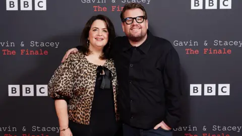 PA Media Ruth Jones and James Corden in front of a black board which has the BBC logo on it (black font of B, B and C with white squares). There is script reading Gavin and Stacey, the finale. And Jones wears a black dress with a leopard print cardigan over the top. Corden has his arm around her, has black specs and a black shirt. They both smile at the camera.