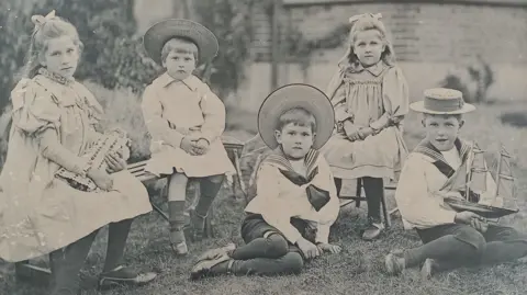 Family of Noel Carrington A black and white image of Noel Carrington and his family. There are five children, all sat down, with two sat on the grass, one holding a boat. Three have straw hats on and two girls are sitting down, with bows in their hair. The girl on the left is holding a doll. They are in front of a building. 