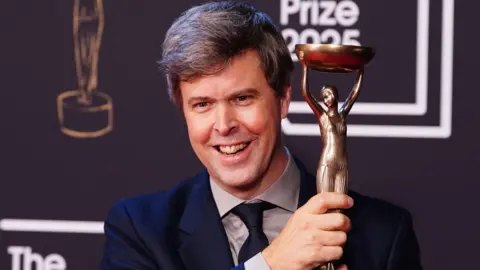 PA Media David Szalay smiling and holding up the Booker Prize trophy