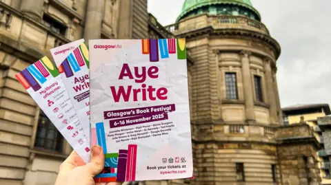 A hand holds the Aye Write book programme with two Aye Write bookmarks. The Mitchell Library is behind them. 
