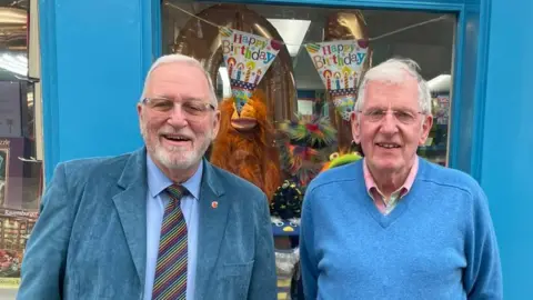 Angela O'Connor Two men standing outside a shop with a blue-painted façade and a sign reading ‘Holman’s’ above the window. The shop window displays books and colourful decorations, including two large orange balloons shaped like animals wearing party hats and a banner that says ‘Happy Birthday.’ 
