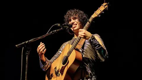 Getty Images Jacob Alon is holding a guitar and singing into a microphone. They are wearing a patterned outfit. 