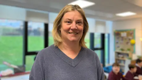 A blonde haired woman with a grey v-necked top smiles at the camera