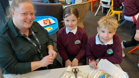 BBC Two children with maroon tops and white collars sit beside a woman in green top with tied back hair. In front of them is a desk with half wrapped books