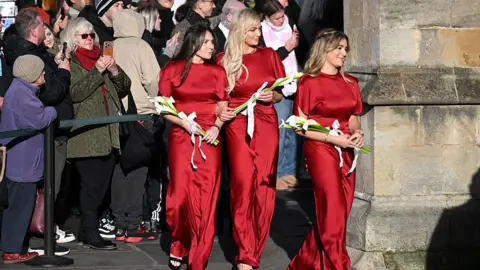Getty Images three women in red full length drsses and carrying what look like bunches of white lillies walk into a church past crowds of onlookers.