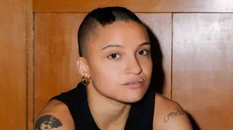 Diana Pfammatter Rene Matić. They have short black hair and are wearing a black vest top. They are sitting in front of a wooden wall. 