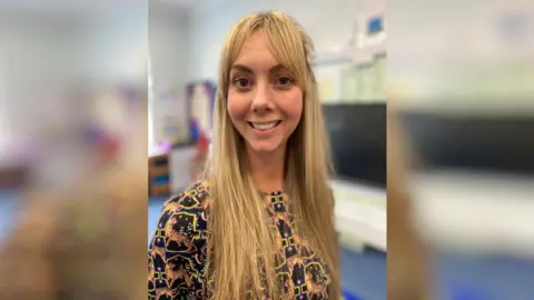 A young woman with blonde hair. She is in a classroom.