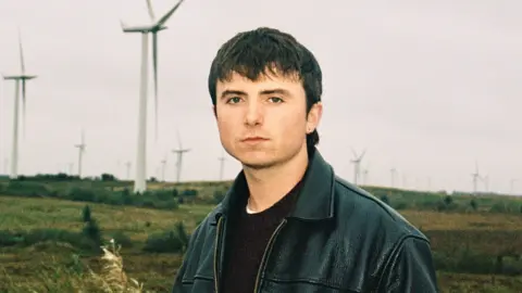 APB PR Shay O'Dowd, a young man wearing a leather jacket and with short dark hair. He is standing outside, with a windfarm visible behind him.