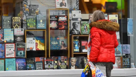 Getty Images A person in a red puffer coat holds shopping bags as they look at book titles displayed in a window of a Waterstones branch in Crewe in 2020.