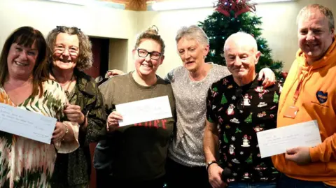 Steve Murray Six people, including Andrew Mears who is second from the right, are standing in a line with their arms slung over each others' shoulders and are smiling at the camera. Three are holding up large cheques. There is a Christmas tree behind them.