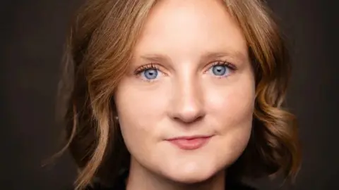 Oliver Doran A woman's face, with short blonde hair and bright blue eyes. She is looking the camera. The background is black. 