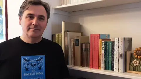 Alma Books Alessandro Gallenzi, a man dressed in a black sweater with short brown hair, holding a book of Dylan Thomas's poetry next to a bookshelf