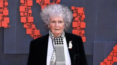 Getty Images Maggi Hambling attends the National Portrait Gallery reopening at National Portrait Gallery on June 20, 2023 in London. She has short, curly grey hair and wears a black jacket with a white top underneath and a black and white scarf with circles on it. 