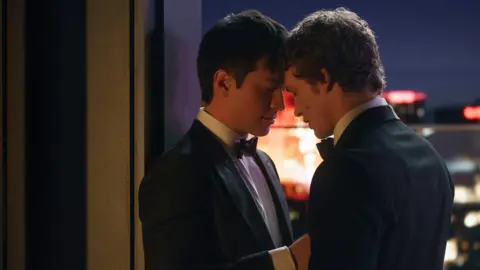 PA Media Heated Rivalry still: Hudson Williams as Shane Hollander and Connor Storrie as Ilya Rozanov. Both men stand with their foreheads pressed together, wearing tuxedos on a rooftop at night.