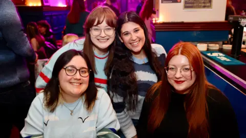 Jonathan Phang Alanna Charlton, Katie Curran, Madison Golan, and Bethan Smith. They are four young women smiling at the camera in a busy pub.