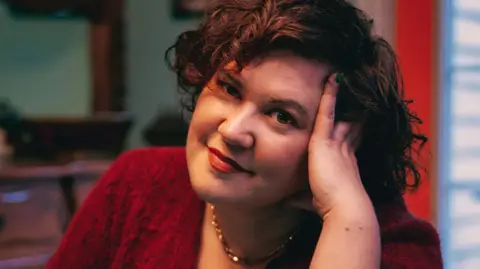 Caleb Latreille Rachel Reid, a woman with short, curly brown hair, leans on her left hand and smiles. She is wearing a dark red top and a gold necklace.