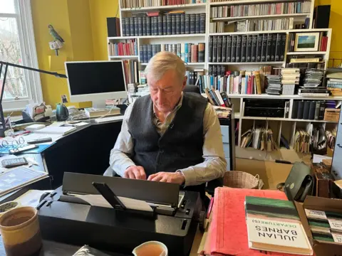 BBC/Roxanne Panthaki Julian Barnes, pale blue shirt and grey sleeveless cardigan, seated at his desk, in his study, typing. Novel Departure(s) on the bottom right, and books on the wall behind