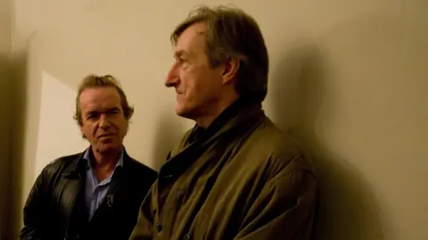 Dafydd Jones Martin Amis on the left -- in a black leather jacket and pale blue shirt - looking at Barnes -- in profile -- in khaki jacket. Both standing against a beige wall
