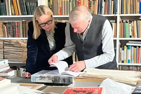 BBC/Roxanne Panthaki Katie Razzall in dark blue velvet jacket, blonde hair and glasses, looking at the notes for Departures that Julian Barnes is showing to her. Both standing. Books behind them & on the table