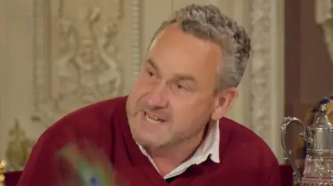 Hugo speaking at the breakfast table with an angry look