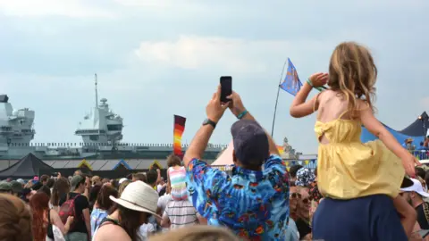 Ben Mitchell/PA Wire A crowd of people in summery clothes look out towards an enormous battleship. On the battleship we can see what appears to be a long row of sailors stood to attention. In the foreground, a little girl in a yellow dress sits upon a man's shoulders while another man in a blue Hawaiian shirt raises his phone in the air to take a picture of the ship. 